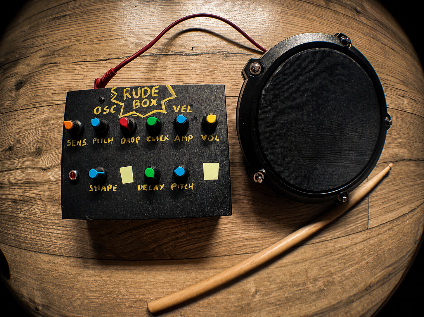 The finished black RudeBox with colorful knobs and hand-lettered yellow labels, beside a drum pad and wooden drumstick.